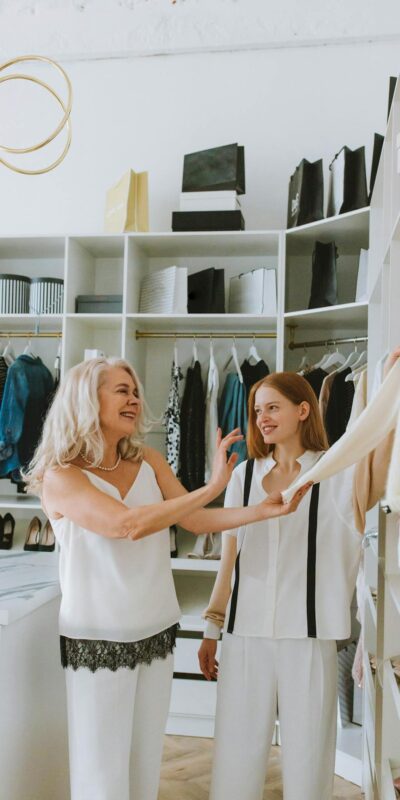 Two women enjoying fashion styling in a chic wardrobe setting with luxury shelves.